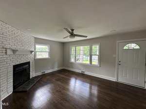 Empty living room with dark hardwood floors, white painted brick fireplace, ceiling fan, three large windows, and a white front door. The walls are light beige, and there is natural light coming in through the windows.