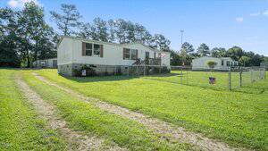 A single-story manufactured home at 160 Lone Wolf Drive in Henderson features white siding, brown shutters, and a fenced yard. A gravel driveway leads to the house, set on a grassy lot with trees and another similar home in the background.