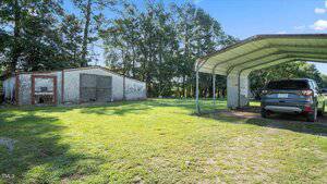 A grassy yard with a weathered shed on the left and a car parked under a metal carport on the right, with trees and a clear blue sky in the background.