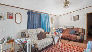 A modest living room with two armchairs covered in blankets, a small table with various items, a fan, and a patterned red rug. The window is covered with blue and navy fabric, and two mirrors and photos hang on the walls.