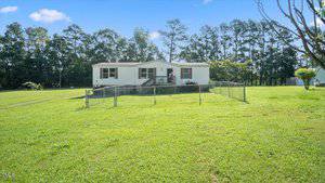 A single-story white manufactured home with brown shutters sits on a grassy lawn, enclosed by a chain-link fence. Trees line the background under a blue sky with scattered clouds.
