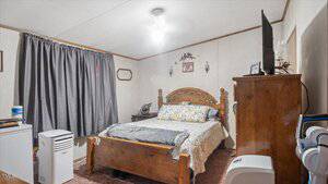 A bedroom with a wooden bed, floral bedding, and matching wooden dresser. A TV sits on the dresser. Gray curtains cover the window. Two portable air conditioners are on the floor along with a mini fridge and lamp.