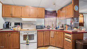 A small kitchen with wooden cabinets, red countertops, a white oven and stove, a coffee maker, a toaster oven, and various kitchen items on the counters. A wall clock and a window with a brown curtain are visible.
