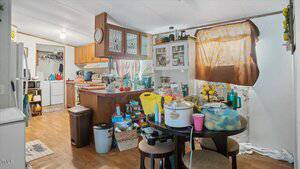 A cluttered kitchen and dining area with wooden cabinets, various food items and containers on the counters and table, a stove, microwave, and a window covered by a brown cloth. The floor is wood, and the room is lit by ceiling lights.