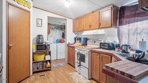 A small kitchen with wooden cabinets, white appliances, and various countertop kitchen gadgets. An open doorway reveals a laundry area with a washer and dryer. Sunlight is partially blocked by a curtain.