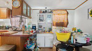 A cluttered kitchen with cabinets, countertops filled with various items, a clock on the wall, and a white hutch in the center. Sunlight filters through brown curtains on the window. A large yellow bin and cooking pots are visible.