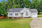 Charming single-story white house in Creedmoor at 2571 Primrose Lane with dark gray roof, attached two-car garage, brick steps to the front door, manicured lawn, trees in the background, and a curved concrete driveway.