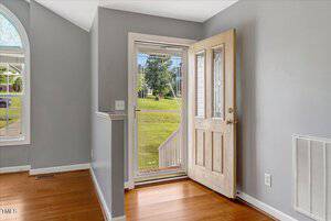 A front door with decorative glass panels is open, revealing a screen door and an outdoor view of a green lawn, stairs, and trees. The room has gray walls, wood flooring, and a large window.