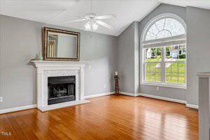 A bright living room with hardwood floors, a white fireplace and mantel, a large arched window, gray walls, a ceiling fan with lights, and a mirror above the mantel.