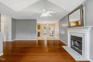 A living room with wood flooring, a white ceiling fan, and a fireplace with a marble surround and a large mirror above it. The space opens to a dining area and kitchen with light cabinetry in the background.