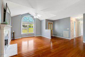 A spacious, empty living room with gray walls, wood floors, a white ceiling fan, a fireplace with a white mantel, and a large arched window allowing natural light inside. A hallway leads to other rooms.