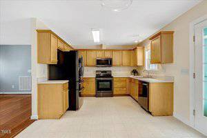 A kitchen with light wood cabinets, black appliances including a refrigerator, oven, microwave, and dishwasher, light countertops, neutral walls, and tile flooring; sunlight enters through a window above the sink.