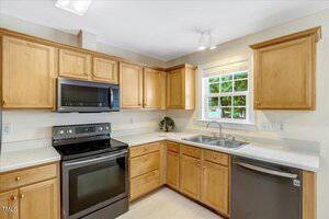 A kitchen with light wood cabinets, white countertops, a black stove with matching microwave, a stainless steel dishwasher, a double sink under a window, and neutral walls. A small plant sits near the window.