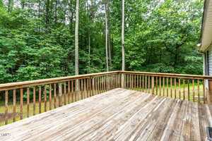 A wooden deck with a simple railing overlooks a yard bordered by tall trees and dense green foliage on a cloudy day.