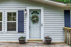 A white house exterior with blue shutters, two windows, and a glass front door. The door has a wreath with the word "Welcome" on it. Two potted plants sit on the wooden porch by the entrance.