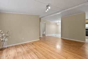 A spacious room with light wood flooring, beige walls, ceiling lights, and a white trim. There is a decorative vase with branches in the corner and an adjoining area with a hanging light fixture visible.