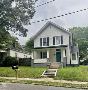 A two-story white house with dark shutters and a covered front porch sits on a grassy lawn with concrete steps leading up to it. A black mailbox is at the curb, and there are trees and neighboring homes nearby.