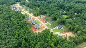 Aerial view of a suburban neighborhood surrounded by dense forest. Several houses are under construction, while some are completed. Roads, cleared lots, and green trees are visible throughout the area.