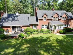A two-story red brick house with black shingles, multiple chimneys, and white-trimmed windows sits among trees, with neatly trimmed bushes and a manicured lawn in front.