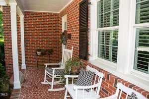 A brick porch with white columns features two white rocking chairs with black-and-white cushions, a potted plant, and a side table. Large windows with shutters are on the right side.