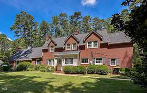 A two-story red brick house with white trim, multiple chimneys, and dormer windows sits in front of a manicured lawn, surrounded by tall pine trees under a blue sky with scattered clouds.