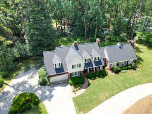 Aerial view of a two-story house with multiple gables, brick and white siding, surrounded by trees and landscaping, with a curved driveway and green lawn in front.