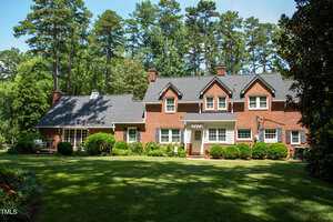 A two-story red brick house with multiple gables, white-trimmed windows, and a manicured lawn, surrounded by tall trees and greenery, under a clear blue sky.
