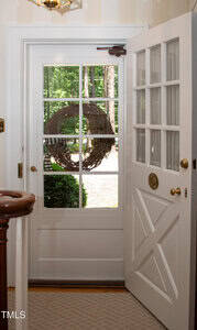 A white wooden door with glass panels is open to the outside. A grapevine wreath hangs on the door. Green trees and shrubs are visible through the glass. Part of a staircase railing appears in the foreground.