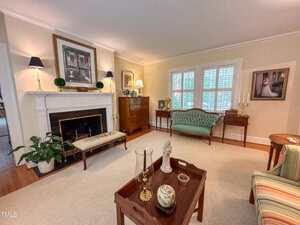 A traditional living room with beige walls, a white fireplace, a green upholstered sofa, wooden furniture, framed artwork, and a coffee table with decorative items. Light streams in through shuttered windows.