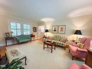 A traditional living room with a cream rug, striped and patterned upholstered furniture, two black-shaded lamps, wall art, and a window with white shutters. Wooden tables and decor items are present throughout the room.