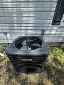 A gray outdoor central air conditioning unit sits on the ground next to a house with white vinyl siding and a black-trimmed window. Grass and some soil are visible around the unit.