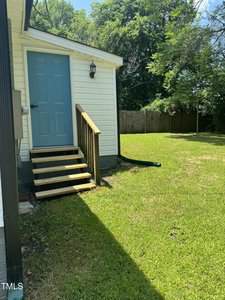 A small set of wooden stairs with a handrail leads to a blue door on a white building. The area is surrounded by green grass and trees, with a wooden fence in the background.