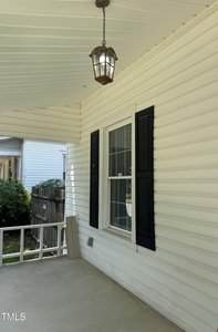 A covered porch with cream-colored siding, a window with black shutters, a hanging lantern light, white railing, and a small portion of a neighboring house visible in the background.