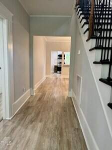 A hallway with light wood flooring, gray walls, white trim, and a staircase with black railings on the right. The hall leads to a room with windows and a view of the kitchen in the background.