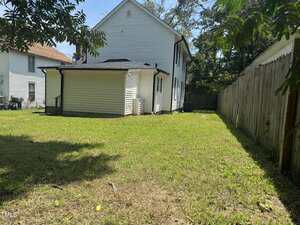 A grassy backyard area bordered by a wooden fence on the right and a white two-story house with black trim in the center. Another house and some trees are visible in the background.