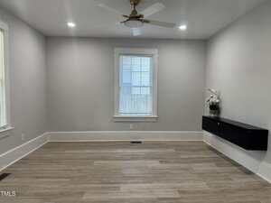 A mostly empty room with light wood flooring, gray walls, a central window with blinds, ceiling fan with light, and a black floating cabinet with a white orchid on the right wall.