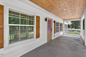 A covered front porch with a wood-paneled ceiling, white siding, three large windows with wood shutters, a beige door with a glass panel, and a concrete floor. A white railing encloses the porch.