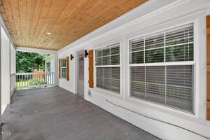 A covered porch with a wooden ceiling, white siding, three large windows with blinds, and a concrete floor. Wall sconces and a railing are visible, with a yard and trees in the background.