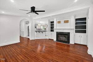 Spacious living room with dark wood floors, white built-in shelves around a fireplace, ceiling fan, and an open view into a white kitchen with a bar counter and pendant lighting.