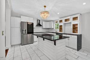 Modern kitchen with white cabinetry, black countertops, stainless steel appliances, center island, a black range hood, glass-front upper cabinets, a farmhouse sink, and a chandelier. The floor has white tiles.