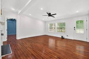 A spacious, empty living room with white walls, a dark ceiling fan, recessed lighting, wood flooring, two large windows, and a glass paneled door. A fireplace and an adjacent room with blue walls are partially visible.