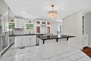 Modern kitchen with white cabinets, black countertops, a central island with shiplap detailing, stainless steel appliances, tile flooring, a chandelier, and windows above the sink allowing natural light.