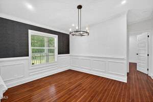 A bright, empty room with white wainscoting, dark accent wall, wooden floor, modern chandelier, and a large window showing green trees outside. Several white doors are visible in the background.