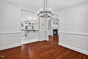 A dining room with white paneled walls, a dark modern chandelier, and polished wood flooring. An arched doorway leads to a kitchen with white cabinetry and island and a living area with built-in shelves and a fireplace.