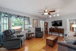 A living room with hardwood floors, two armchairs, a sofa, a wooden coffee table, a sideboard with a clock and TV on the wall, and large windows with sheer curtains letting in daylight.