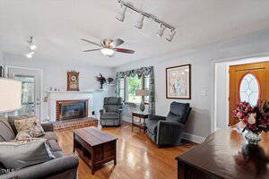 A living room with hardwood floors, gray upholstered seating, a wooden coffee table, and fireplace. Track and ceiling lights, a ceiling fan, artwork, and decorative items are visible. A door with an oval window is at right.