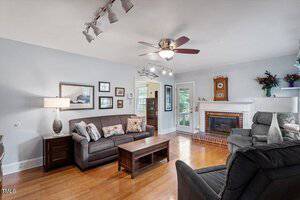 A living room with wooden floors, a gray sofa, a coffee table, and an armchair. There is a ceiling fan with lights, a fireplace with a mantle, wall art, and a window and door letting in natural light.