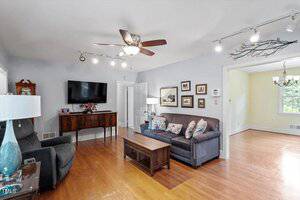 A living room with hardwood floors, a gray sofa, armchair, coffee table, TV mounted on the wall, sideboard with decor, framed art on the walls, ceiling fan, and track lighting. An adjacent room is visible.