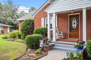 A red brick house with a small covered front porch, a white rocking chair, potted flowers, and a wooden front door with an oval glass panel. Shrubs and a flower bed line the walkway leading to the steps.