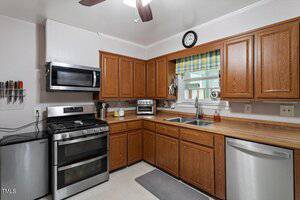 A kitchen with wooden cabinets, stainless steel appliances including a stove, microwave, and dishwasher, a double sink under a window with a plaid valance, and utensils on the counter and wall.